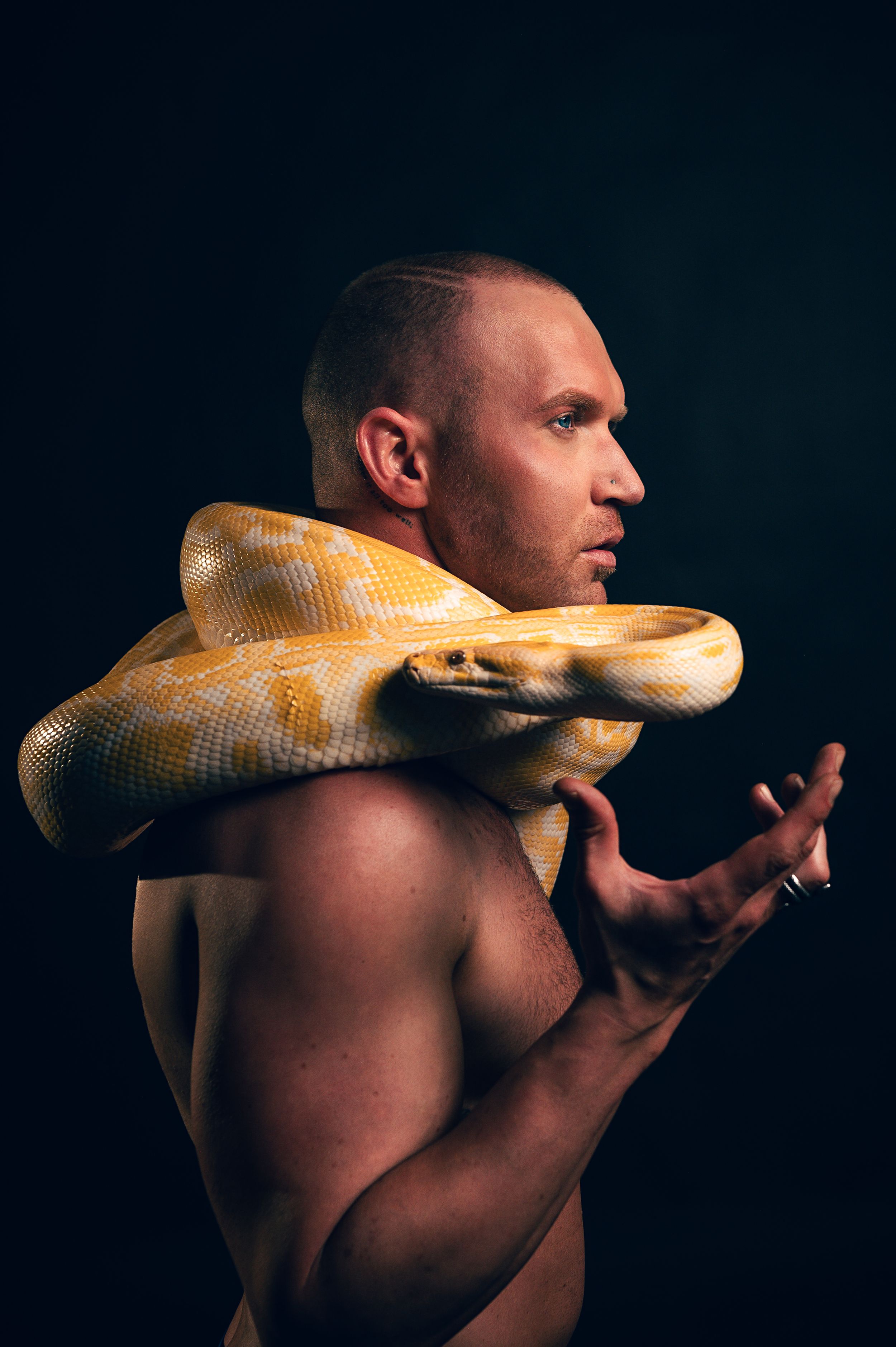 Shirtless muscular man holding a large albino yellow python snake draped around his shoulders on dark background.