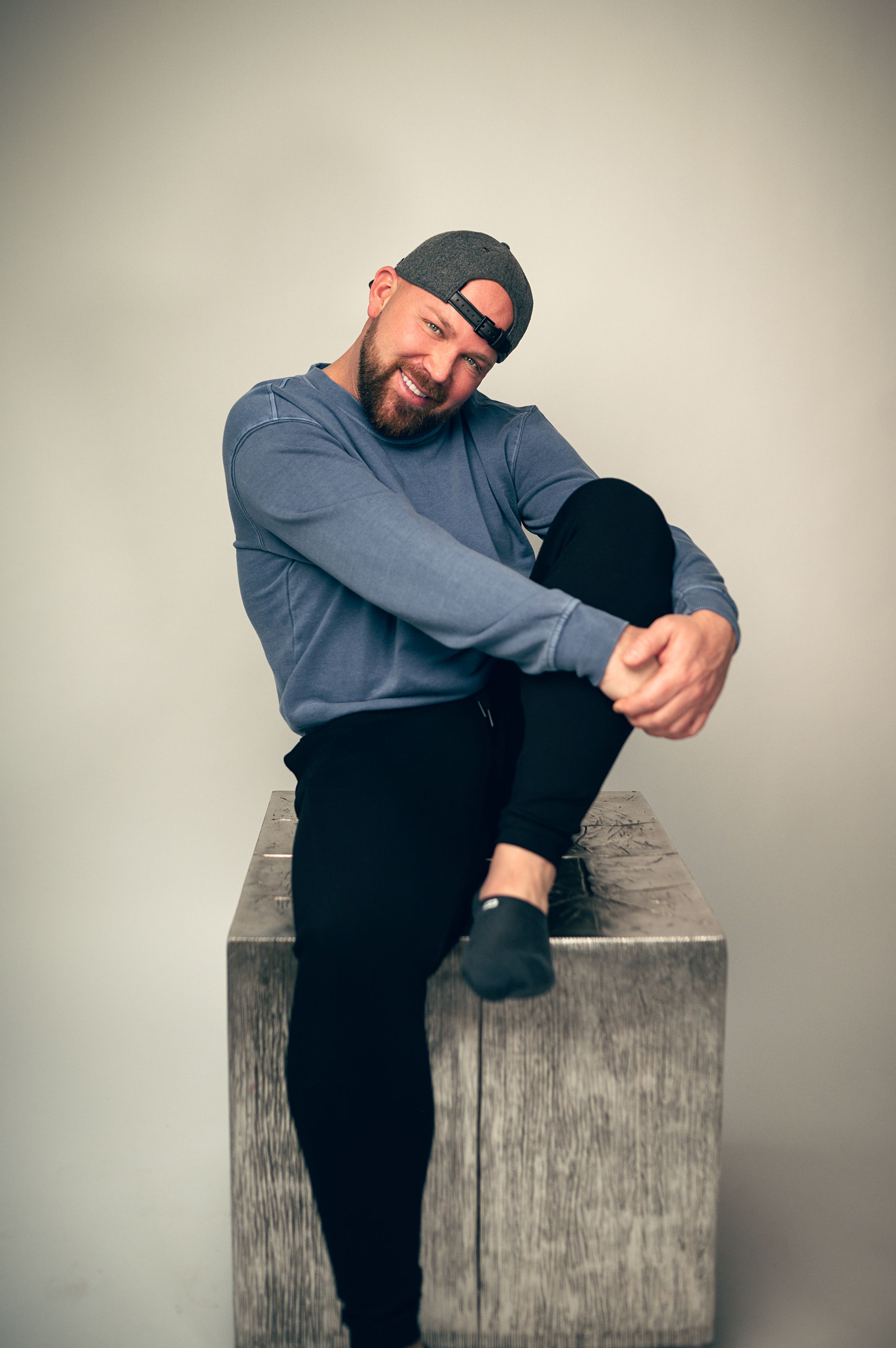 Smiling bearded man in a gray sweatshirt and backward cap sitting relaxed on a wooden block in a studio portrait.