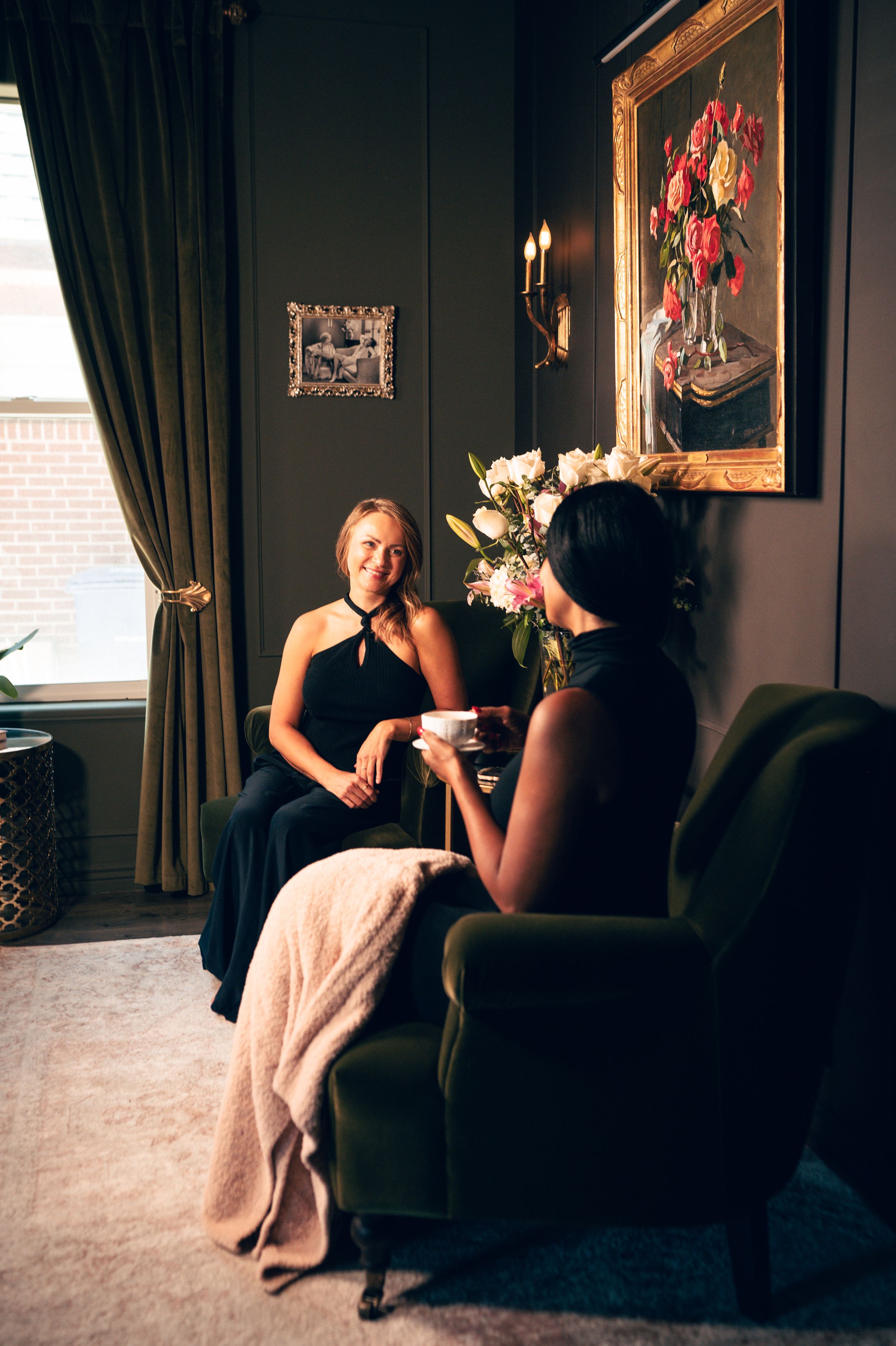 Two women in black outfits having a relaxed conversation in an elegantly decorated dark-toned living room.