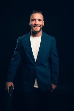 Smiling bearded man in navy blazer and white tee against dark background.