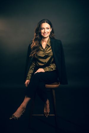 Woman in olive blouse and black blazer sitting on stool against dark background.