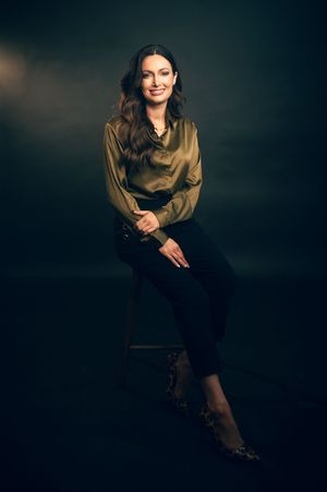 Smiling woman in gold satin blouse and black pants seated on stool in dark studio.
