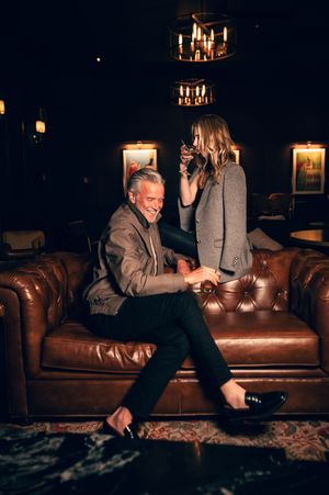 Silver-haired man and blonde woman laughing together on leather sofa in warmly lit elegant lounge bar.