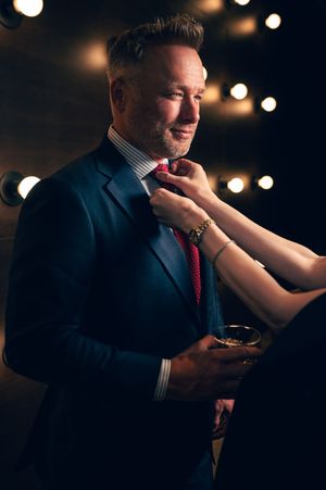 Smiling man in navy suit having red tie adjusted while holding whiskey glass in moody lit setting.