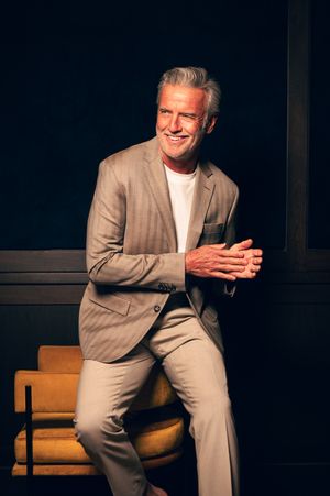 Smiling middle-aged man in beige suit sitting relaxed against dark background.