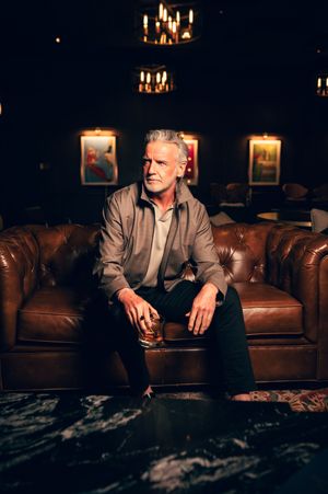 Distinguished silver-haired man in casual jacket sitting on leather sofa holding drink in dark upscale lounge.