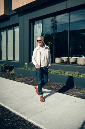 Stylish silver-haired man in white jacket and jeans walking confidently on sunny urban sidewalk wearing sunglasses.