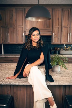 Confident woman in black top and white pants seated at kitchen counter.