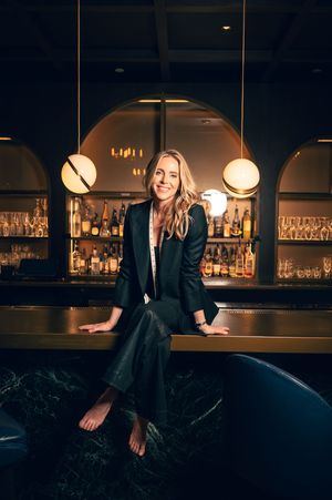Smiling blonde woman in black blazer sitting barefoot on bar counter in elegant dimly lit cocktail bar.