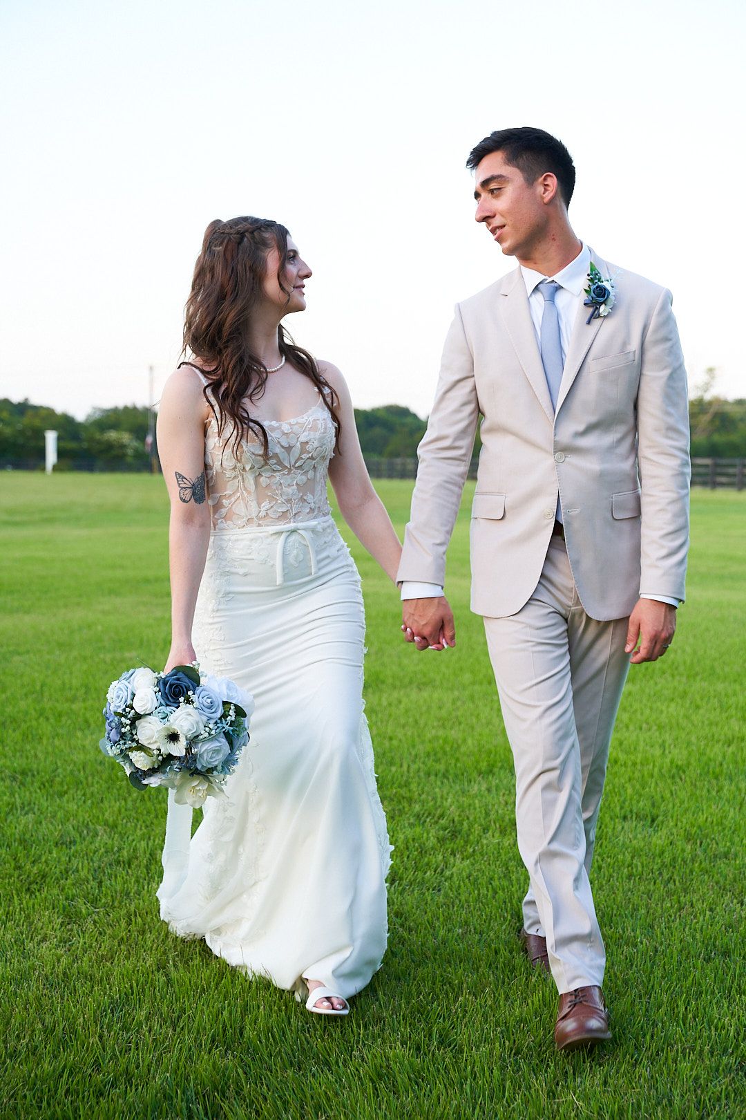 A bride and groom walk hand in hand through a lush green field wearing white and tan outfits while holding a blue bouquet.