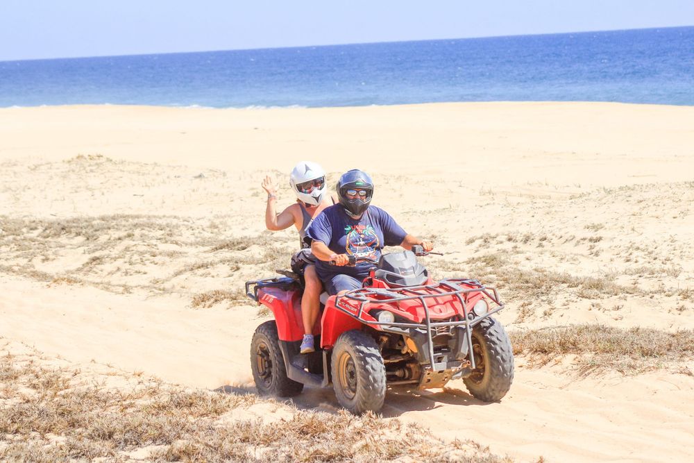 Someone rides a red ATV on a sandy beach with blue ocean waters in the background.