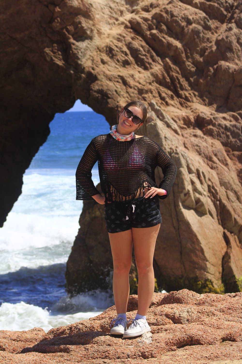 Tourist poses in front of natural rock arch formation with blue ocean waves crashing in background at scenic coastline.
