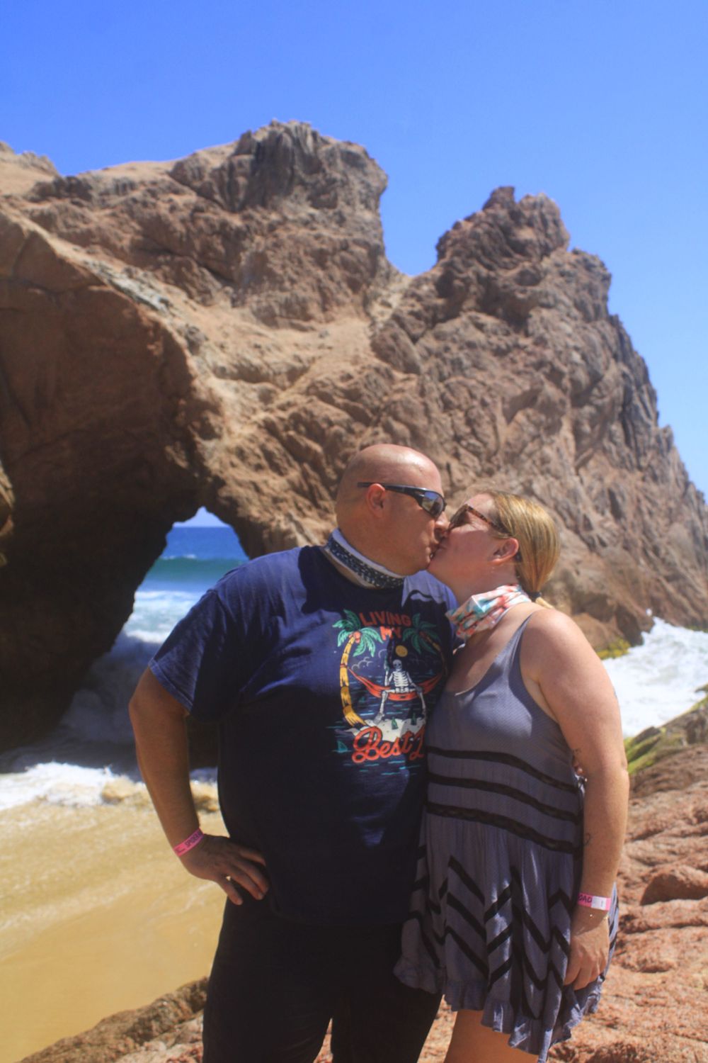 A couple embraces on a rocky beach with waves crashing behind them and tall cliffs in the background.