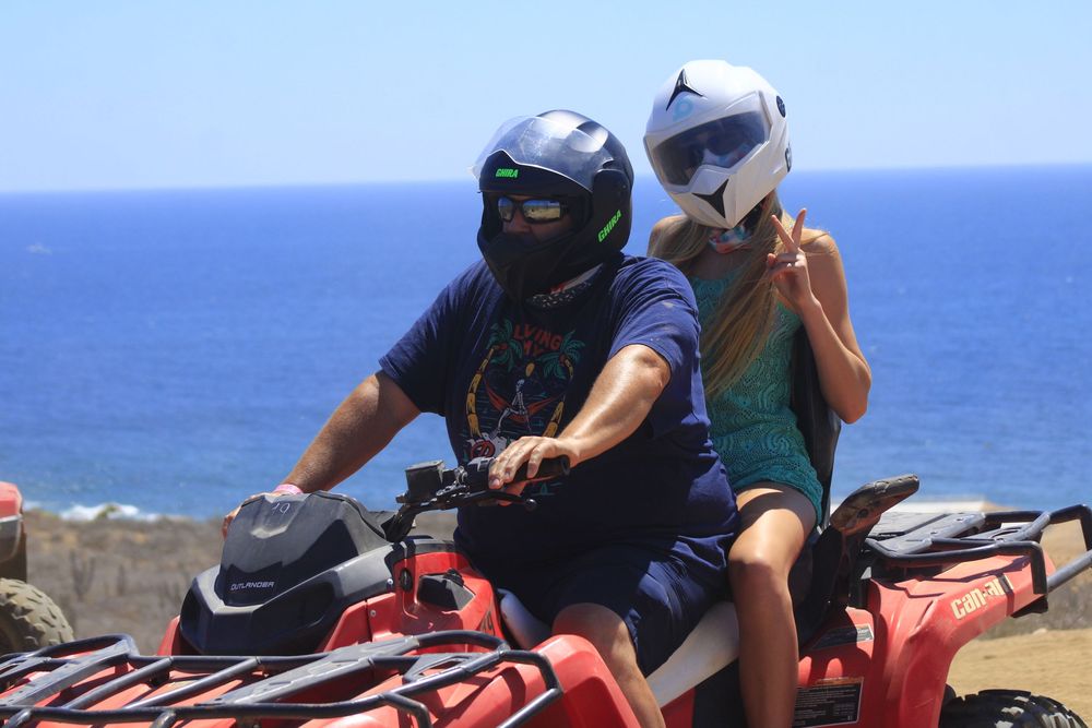 Two riders on a red ATV enjoying an oceanside adventure on a sunny day with blue water in the background.