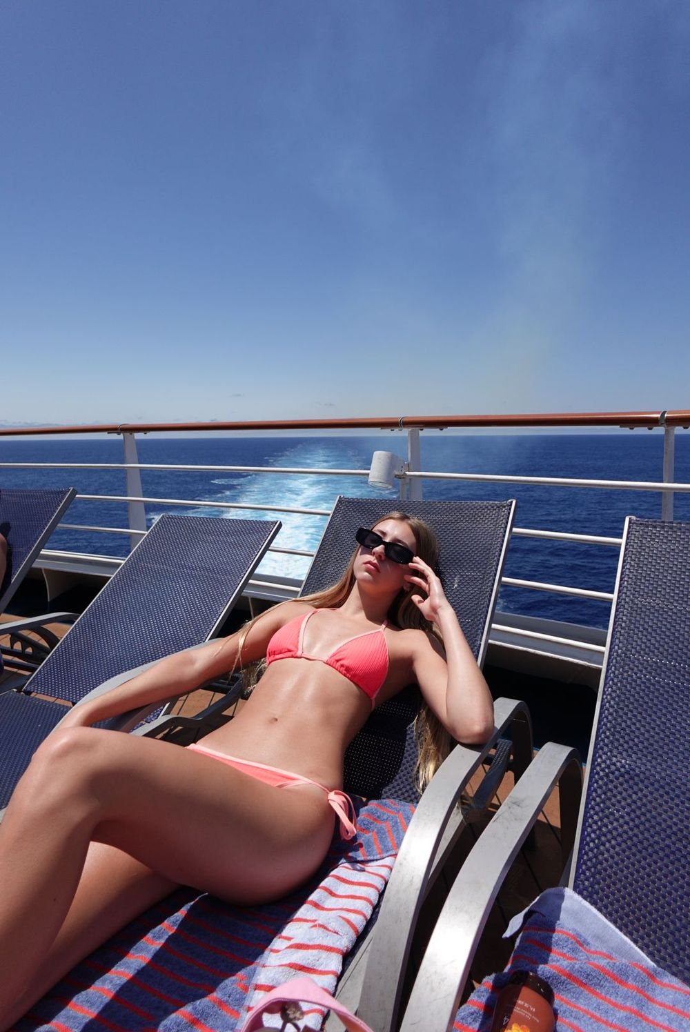Person in coral bikini relaxing on lounge chair with solar panels visible against bright blue sky at beachside location.