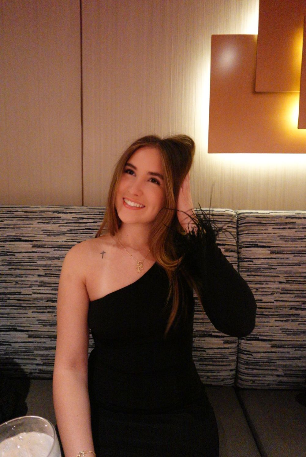 A person in an elegant one-shoulder black dress poses at a stylish restaurant with ambient lighting.