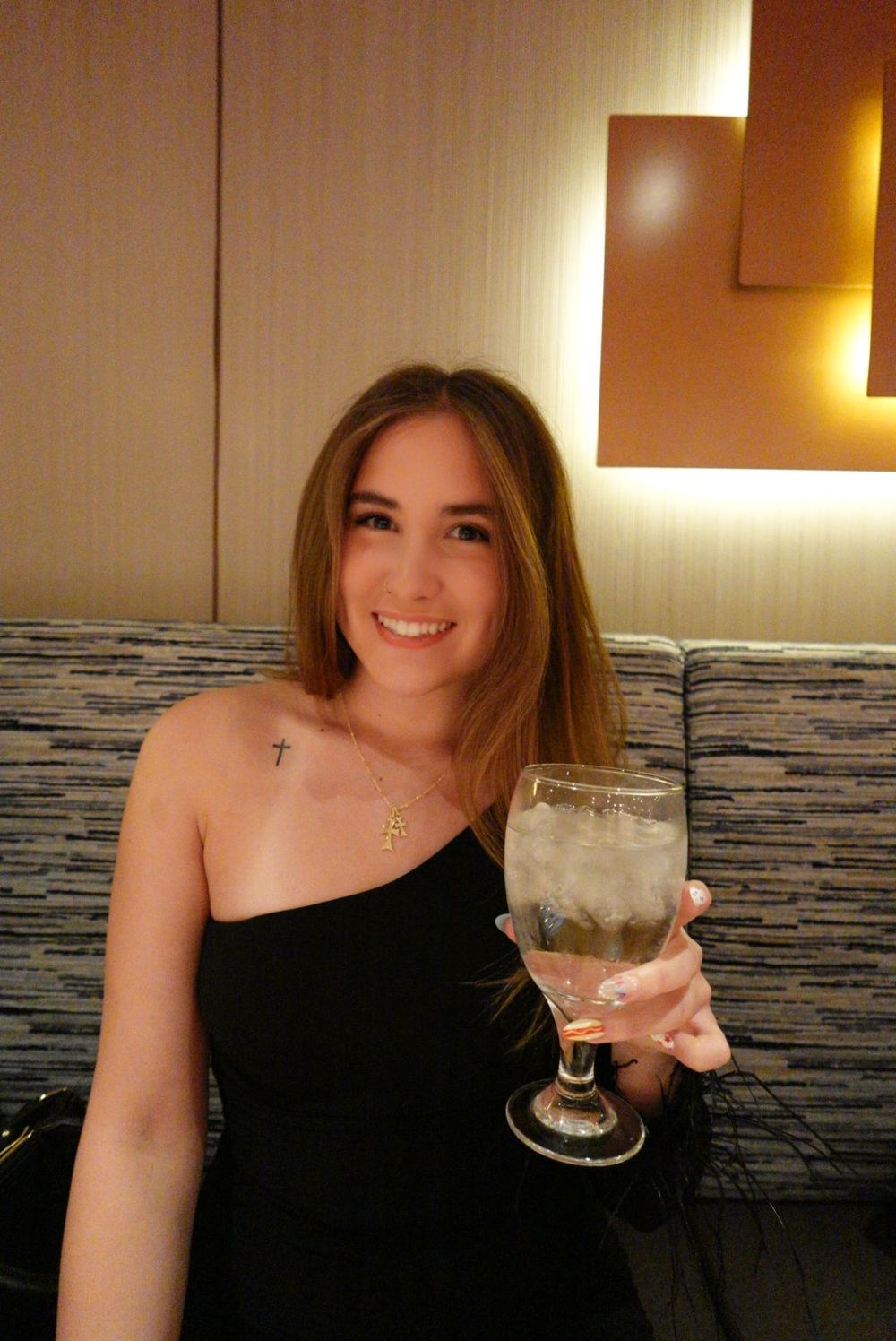 A smiling young lady in a black one-shoulder dress holds up a frosted glass of cocktail in an ambient-lit setting.