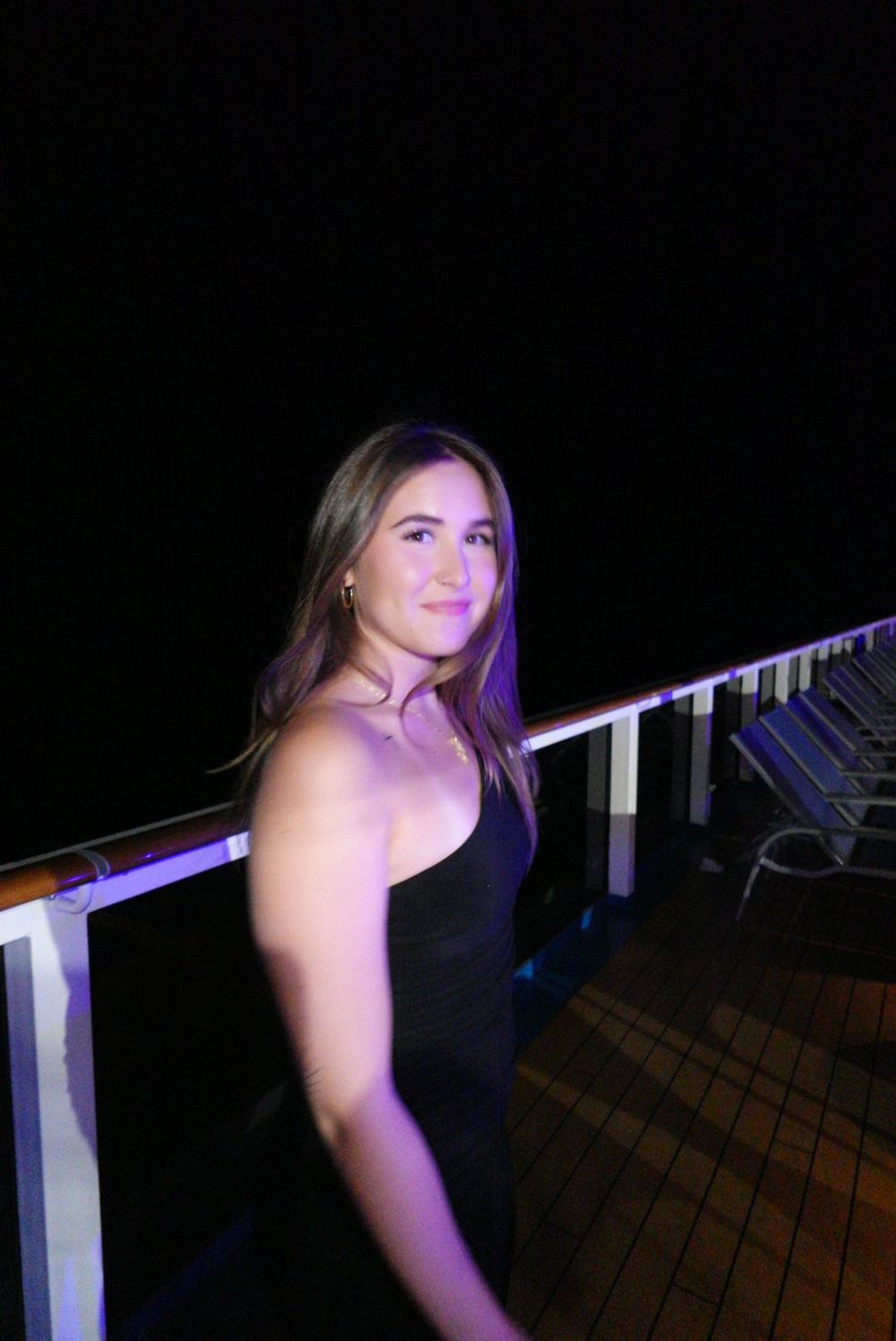 Someone in a black dress poses on a cruise ship deck at night with purple lighting.