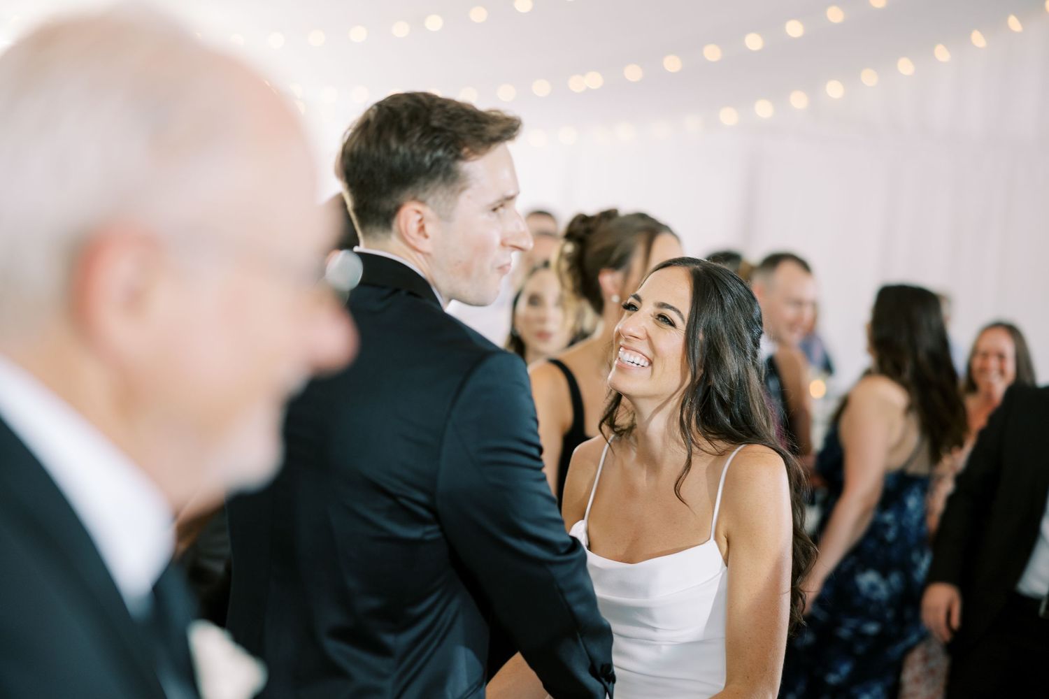 A joyous newlywed couple shares their first dance at a wedding reception as guests look on with lights twinkling overhead.