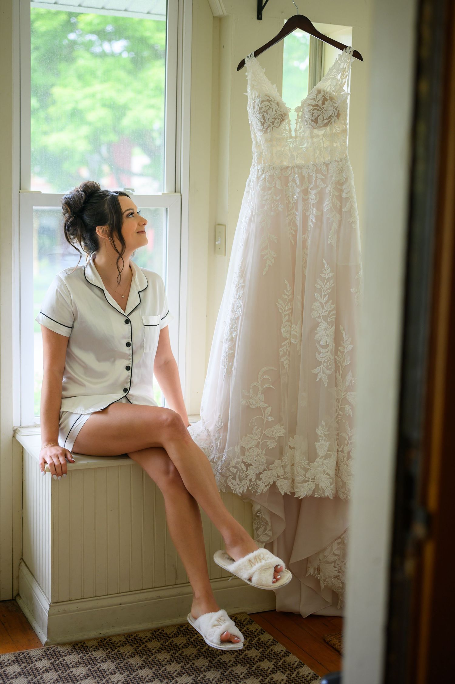 A wedding dress hangs near a window while someone relaxes in casual white attire before the ceremony.