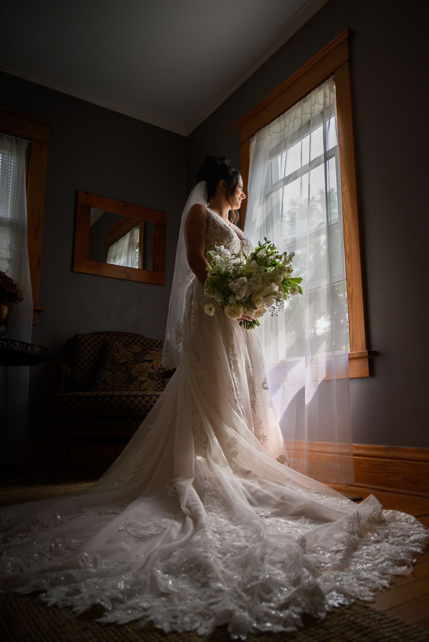 A flowing white wedding gown with delicate lace details is beautifully backlit by natural window light in an indoor setting.