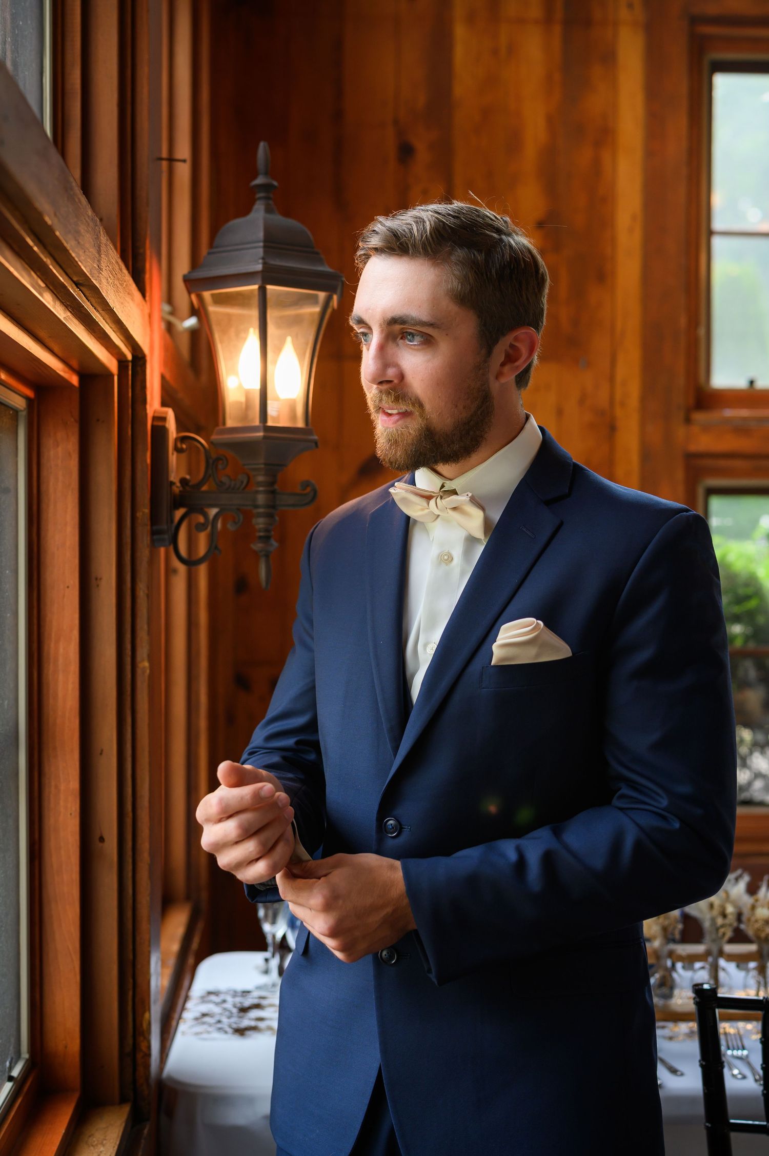 A well-dressed figure in a navy suit with cream bowtie stands by a window with vintage wall lamp.
