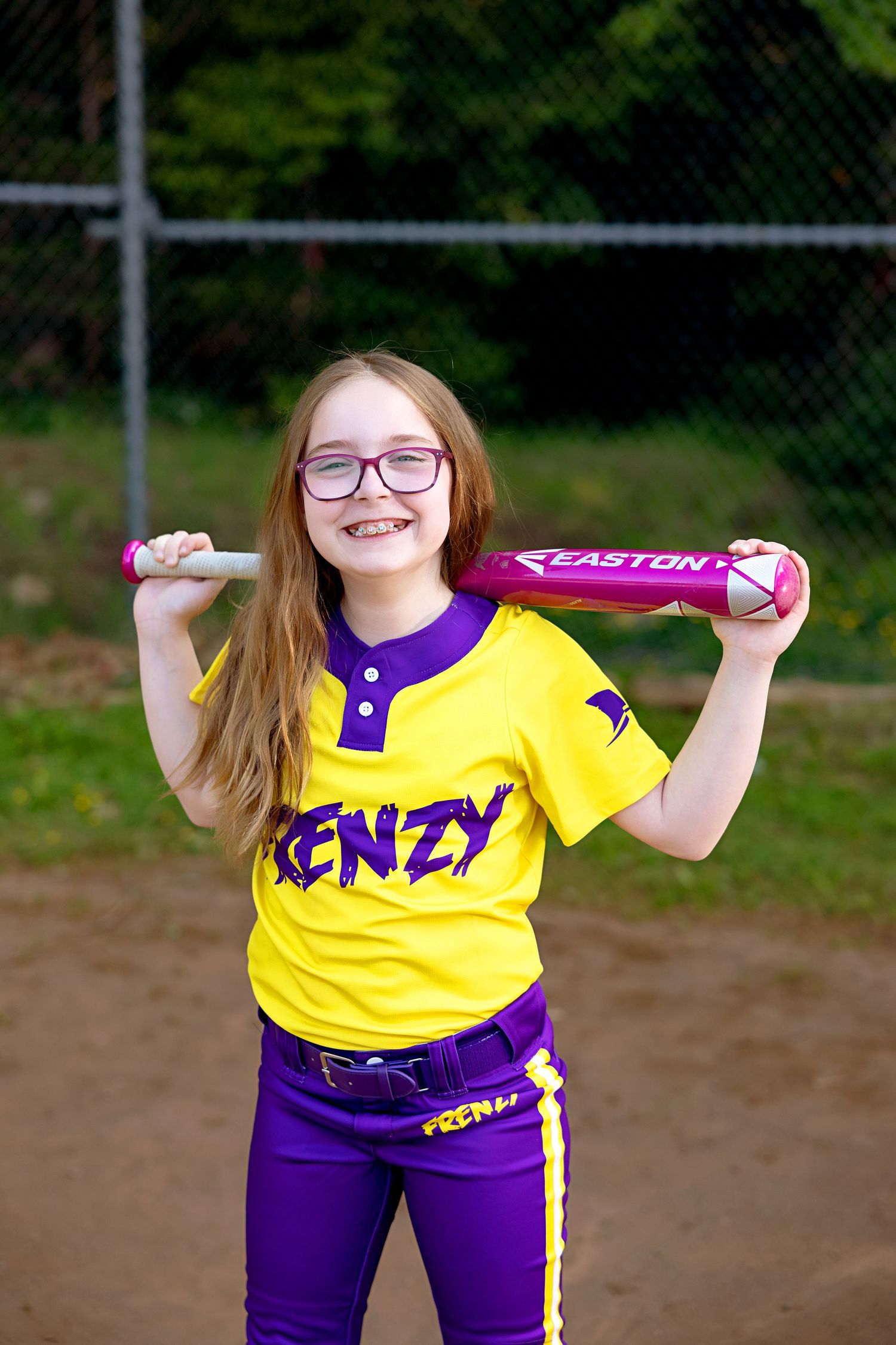 Softball player in yellow and purple Frenz uniform tosses yellow ball on dirt field with chain link fence in background.