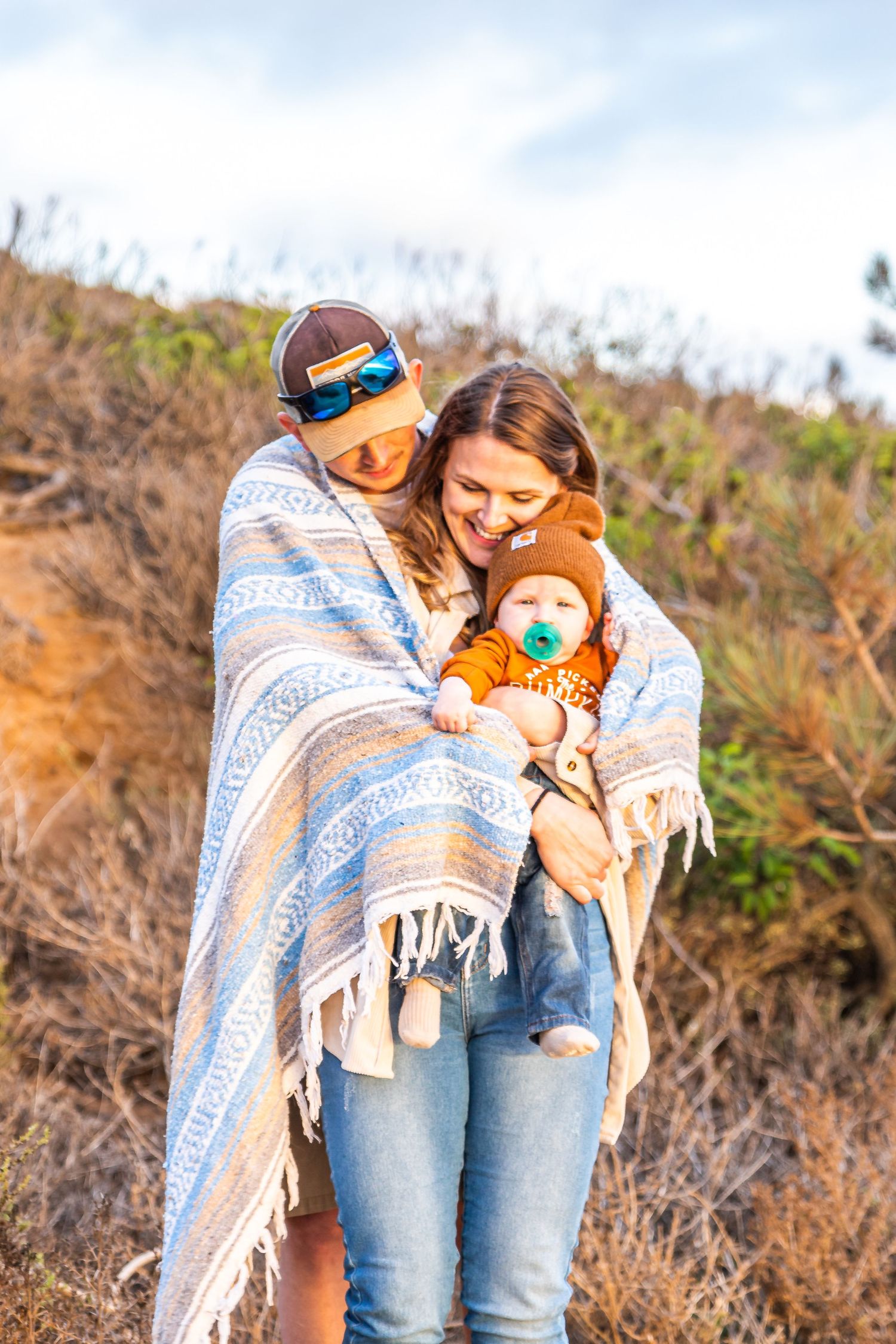 People wrapped in a blue and white blanket share a joyful moment outdoors with a stuffed animal on a sunny day.