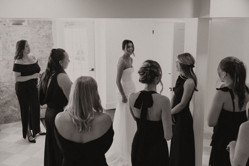 Wedding party members in black attire gathered in a white-walled room.