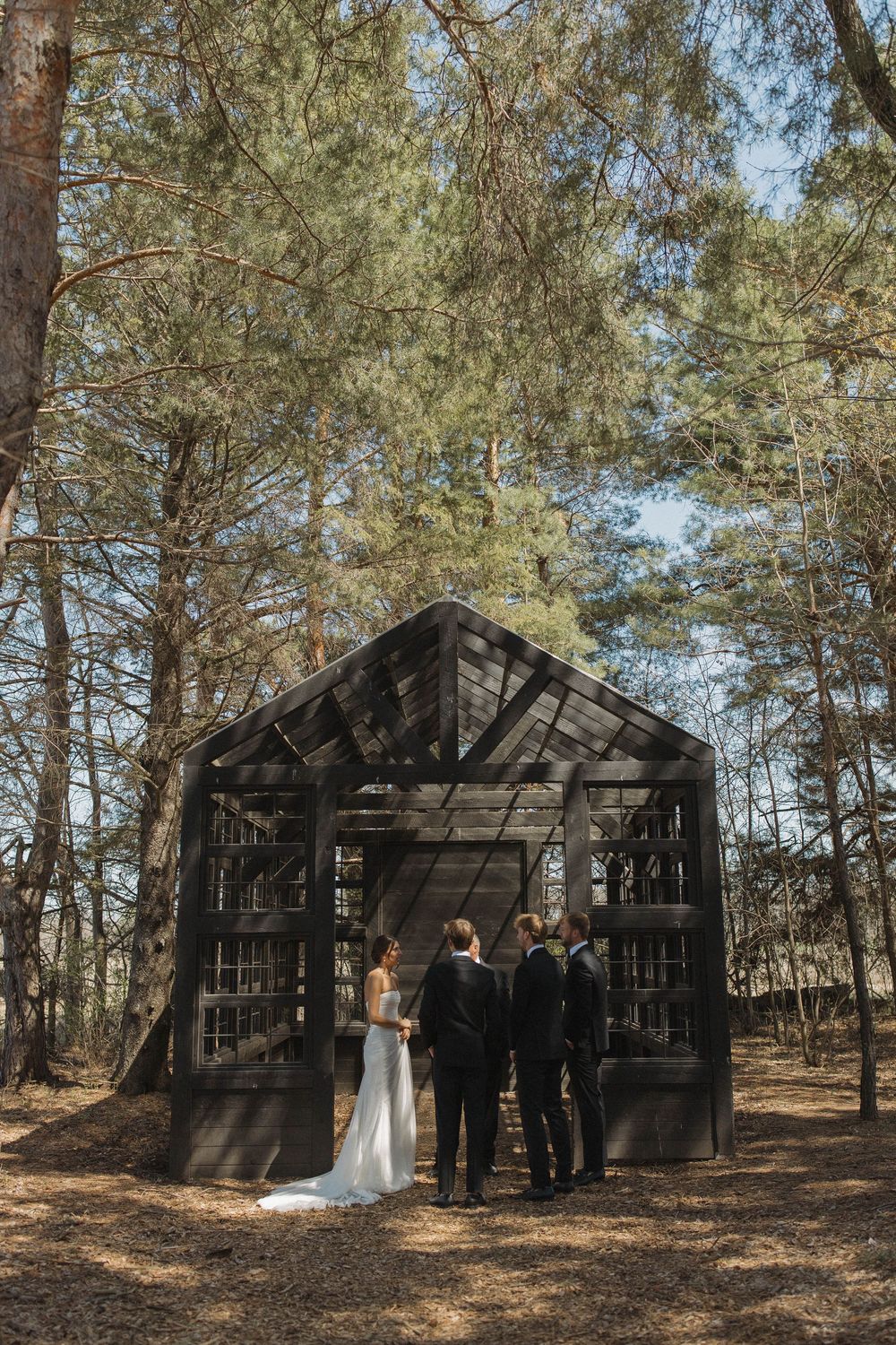 Wedding ceremony taking place in front of rustic wooden structures in a pine forest.