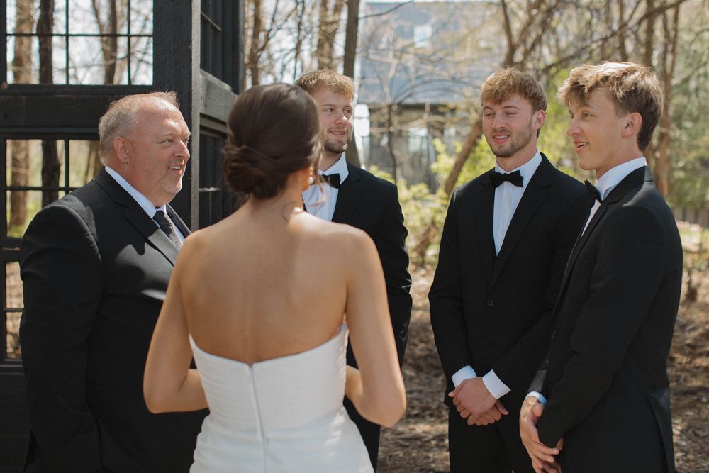Wedding party gathered outside rustic wooden structures for formal photographs.