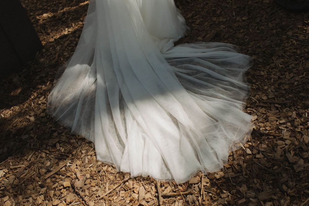 Flowing white wedding dress train spread out on forest ground in natural lighting.