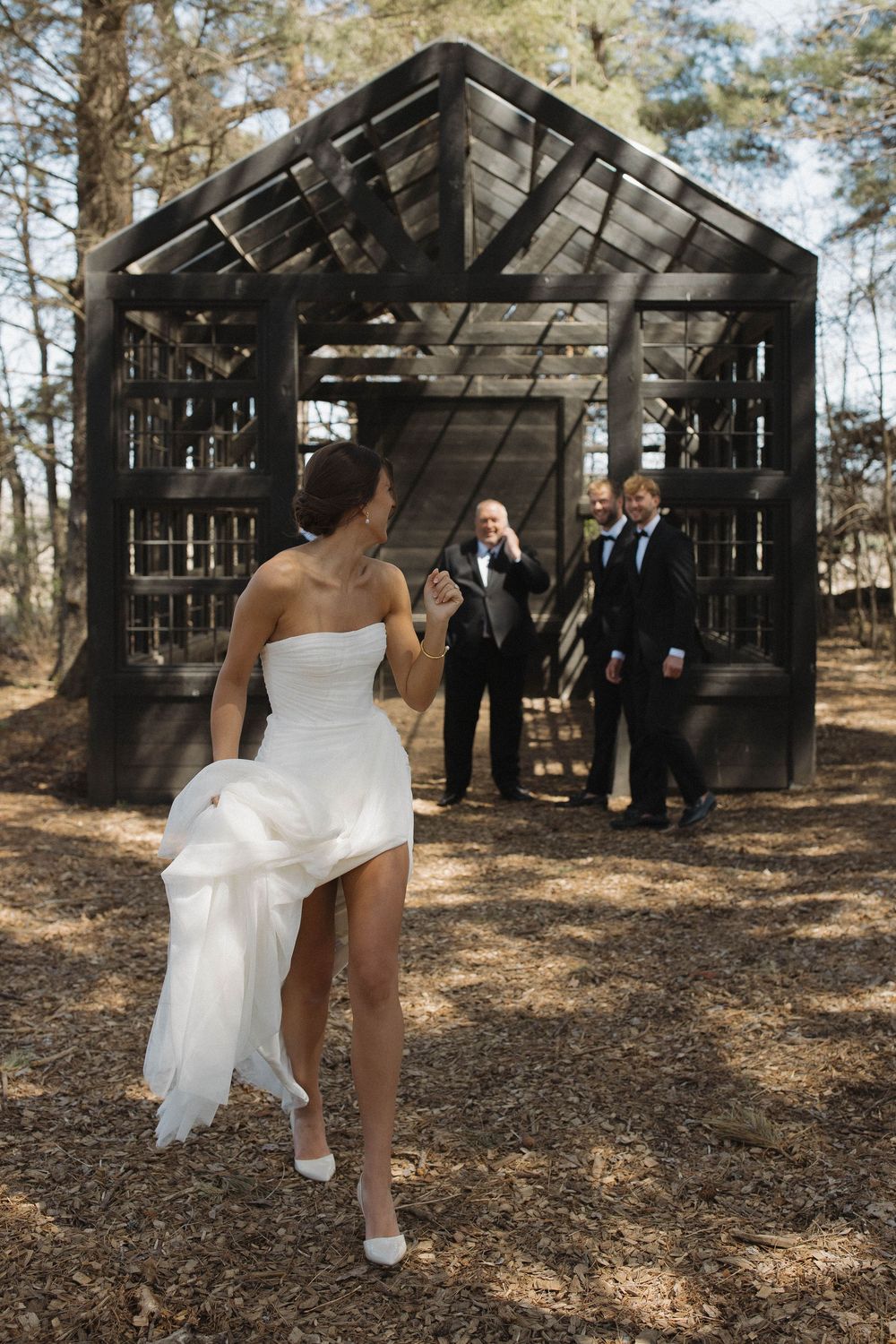Bride in white dress showing off her dress near rustic wooden structures in a forest setting.