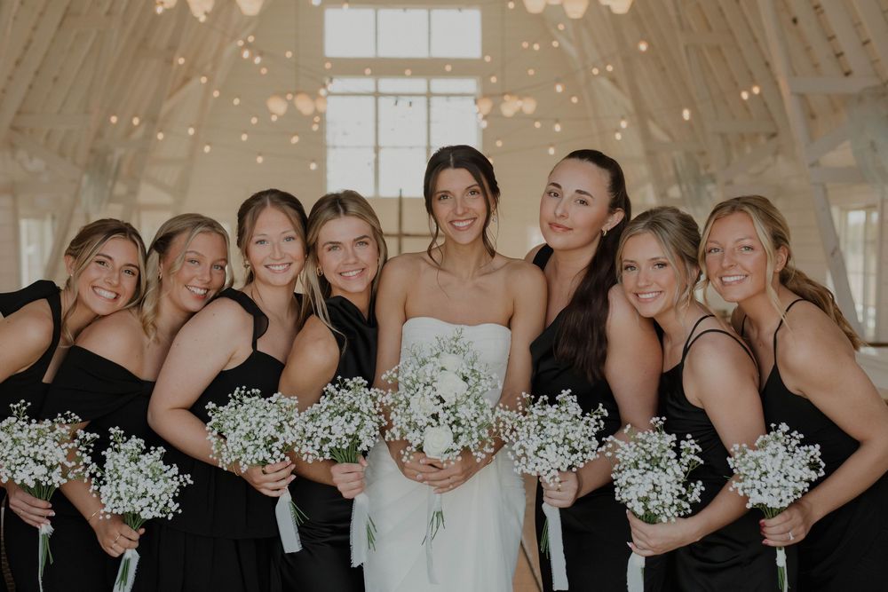 Bridesmaids in black dresses share a candid moment while holding white baby's breath bouquets at an indoor venue.