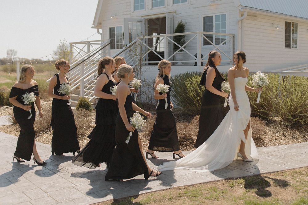 Wedding party sits casually outside a white farmhouse-style venue on a sunny day.