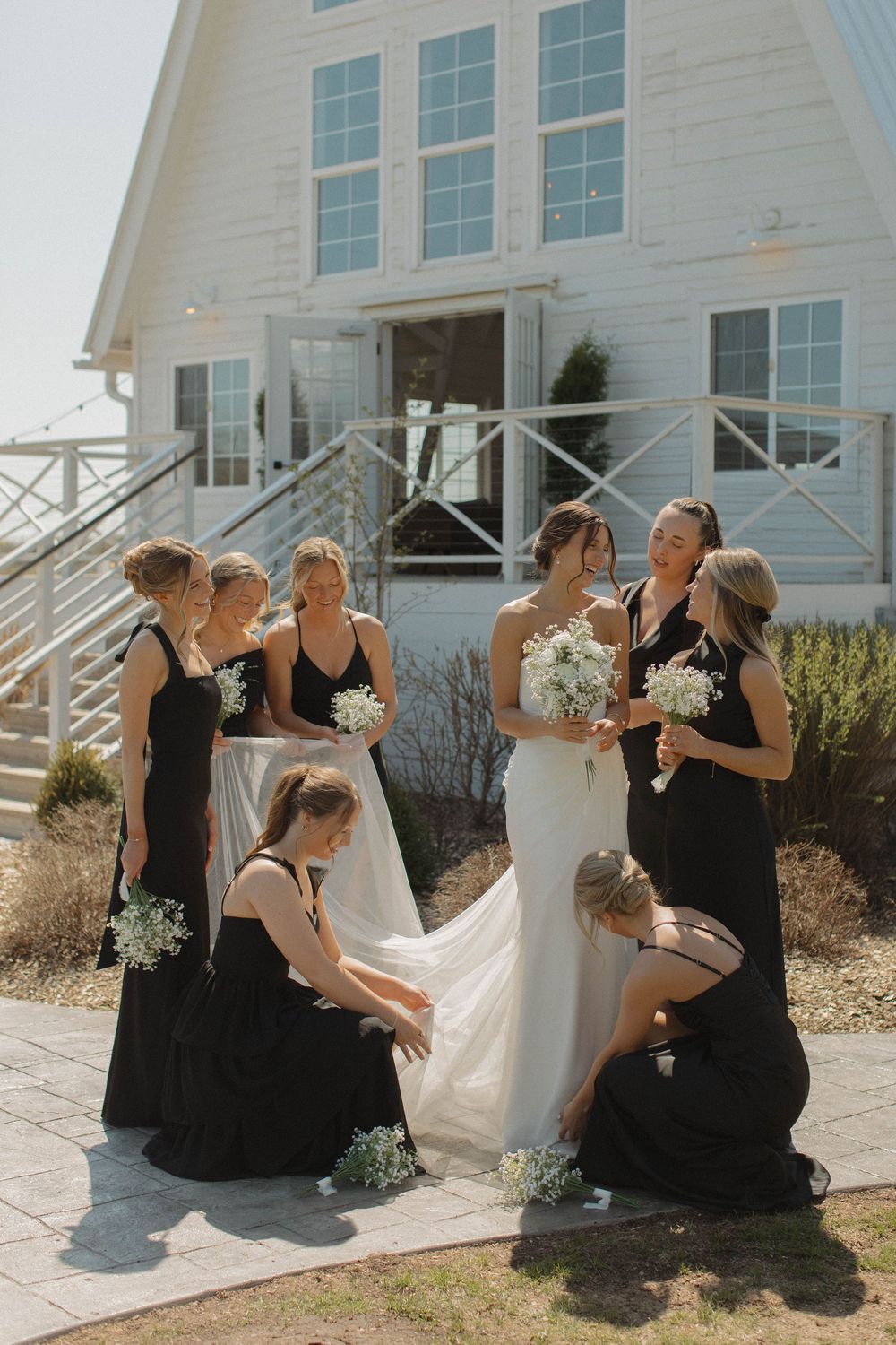 Bridesmaids in black dresses share laughs with bride outside white barn venue.