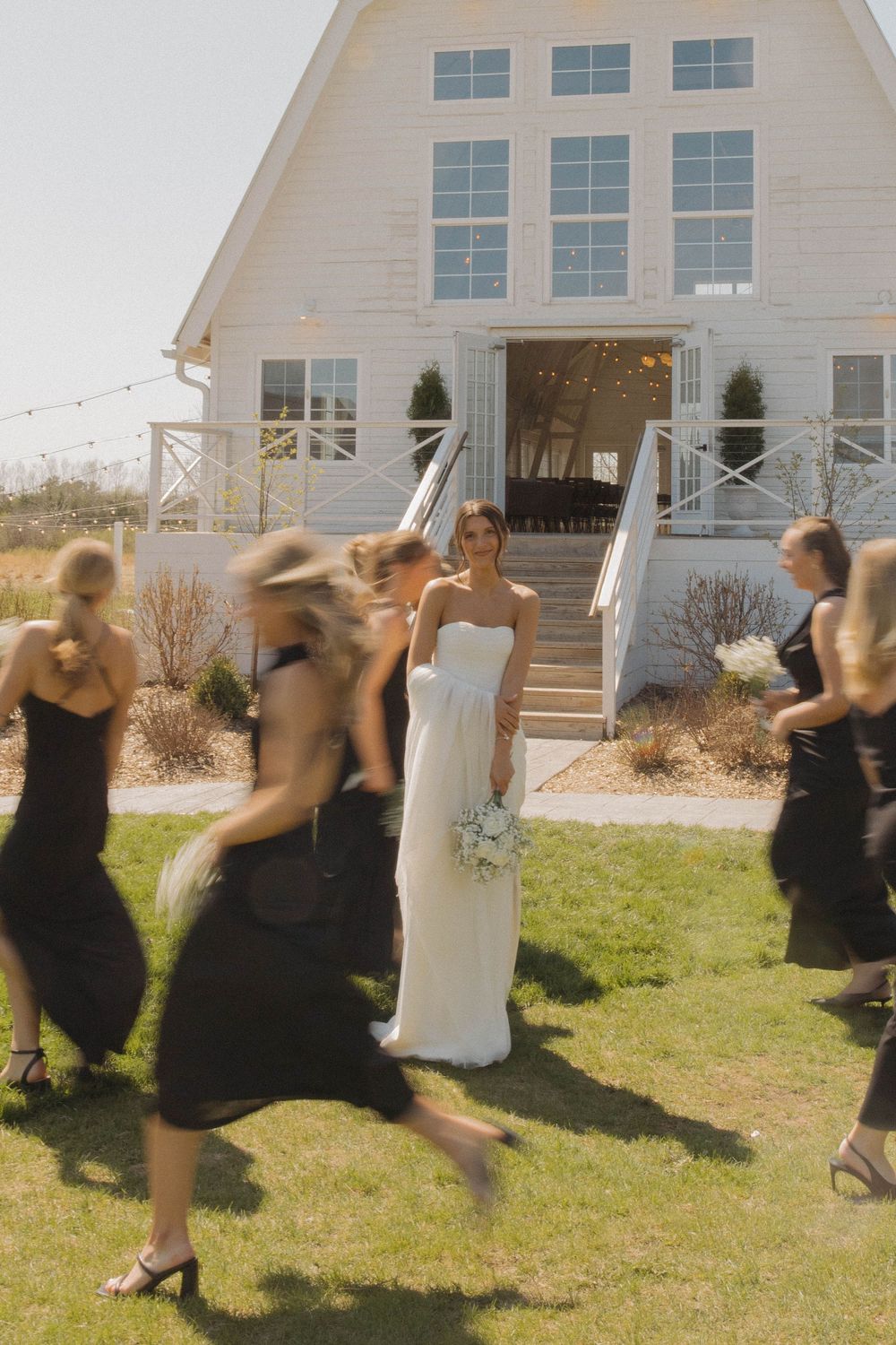 Wedding party dances and jumps playfully on grass outside white farmhouse building.