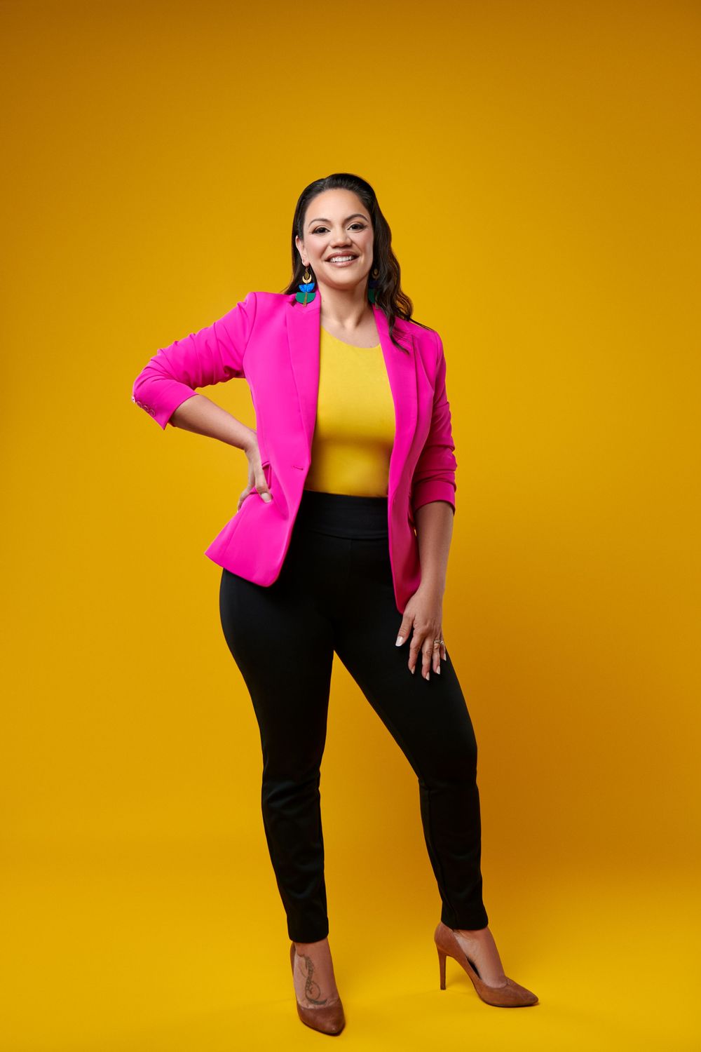 Someone wearing a hot pink blazer and black pants poses confidently against a vibrant yellow backdrop. Gricel's Photography