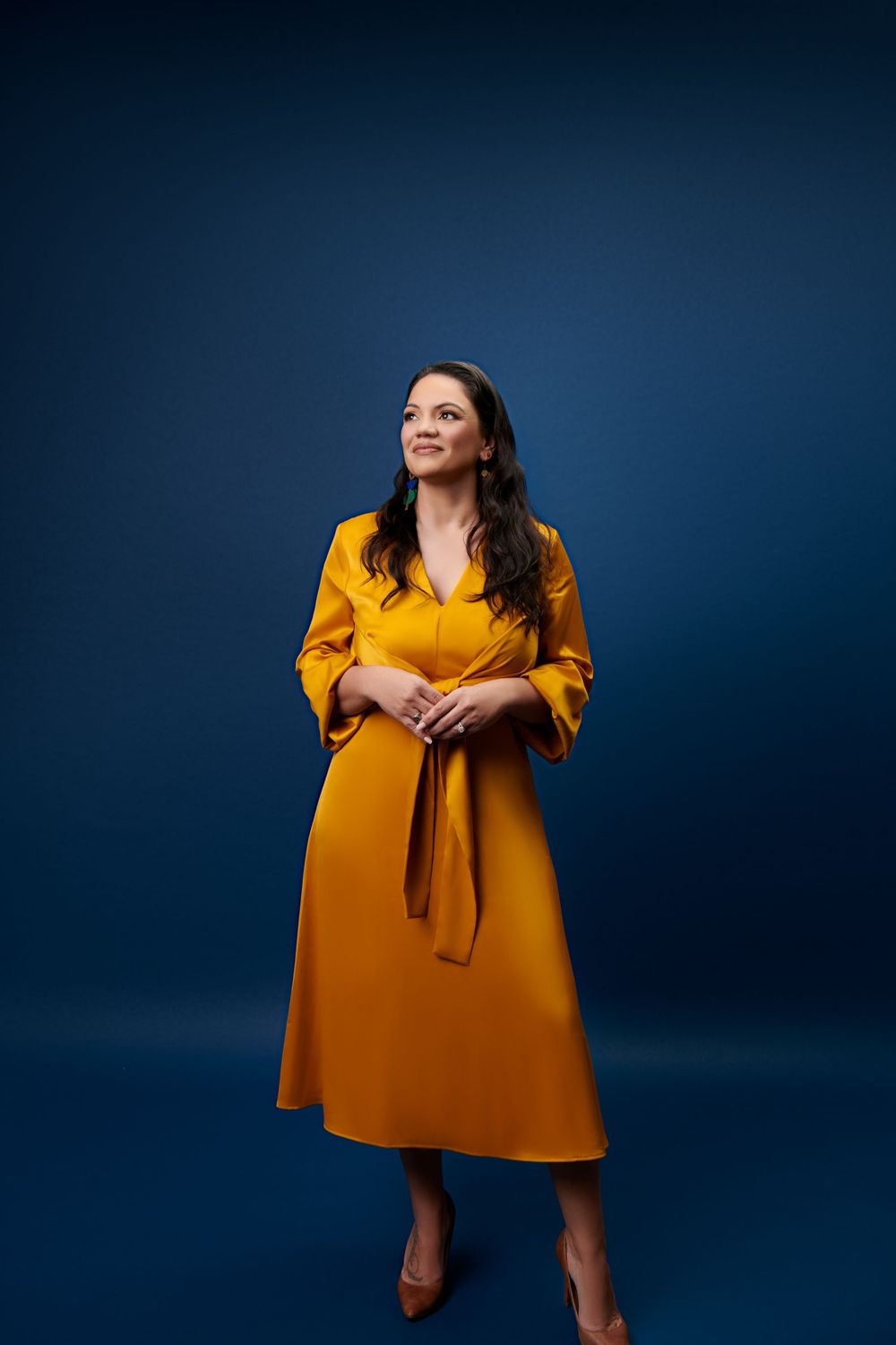A person in a vibrant mustard yellow midi dress with three-quarter sleeves poses against a dark blue studio backdrop.