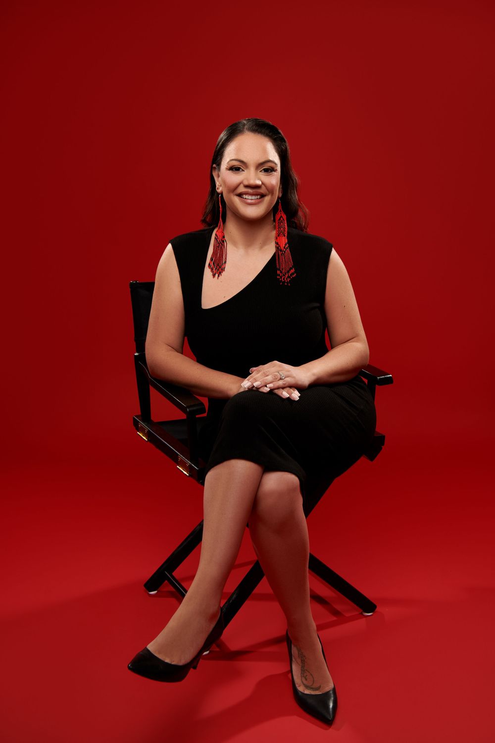 A person wearing a black dress sits in a director's chair against a vibrant red backdrop.