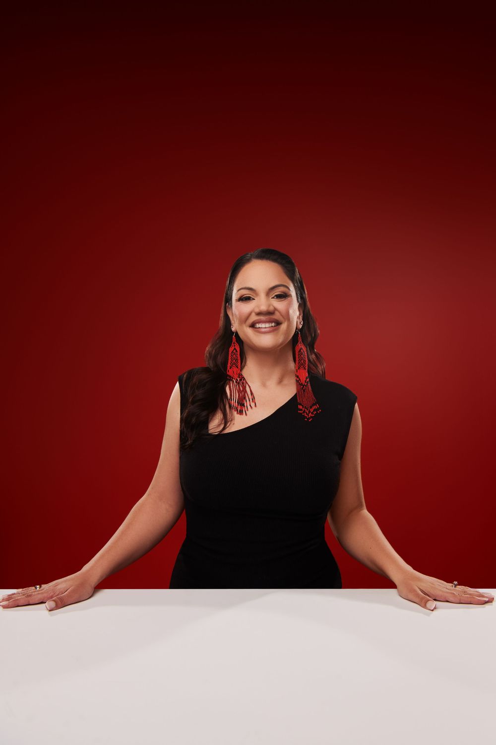 Someone in a black sleeveless top poses against a red background while leaning on a white surface with a warm smile.