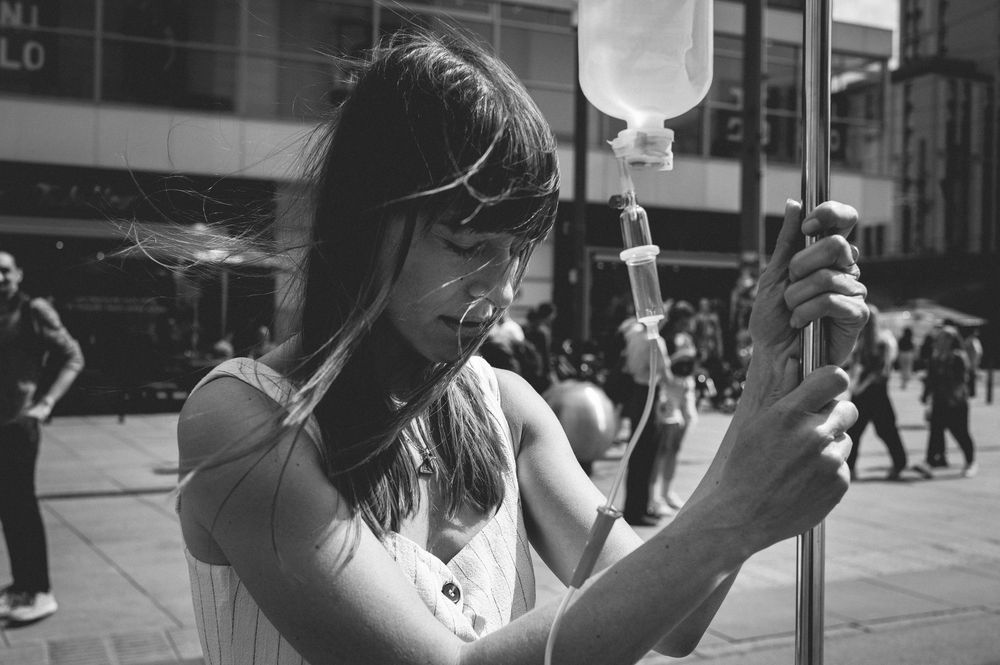 A black and white grayscale photo shows someone on a city street holding an IV drip bag in their hand.