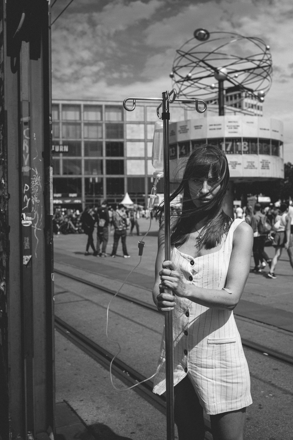 Woman in a light-colored outfit stands outdoors holding a device on what appears to be a city street or platform.