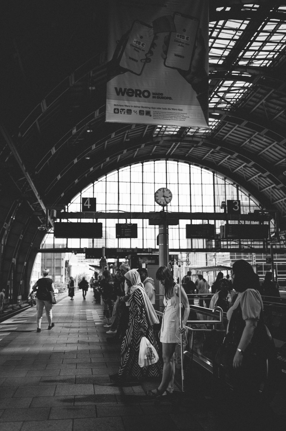 Black and white panoramic view of silhouetted travelers inside a vintage railway station concourse with arched ceiling panels.