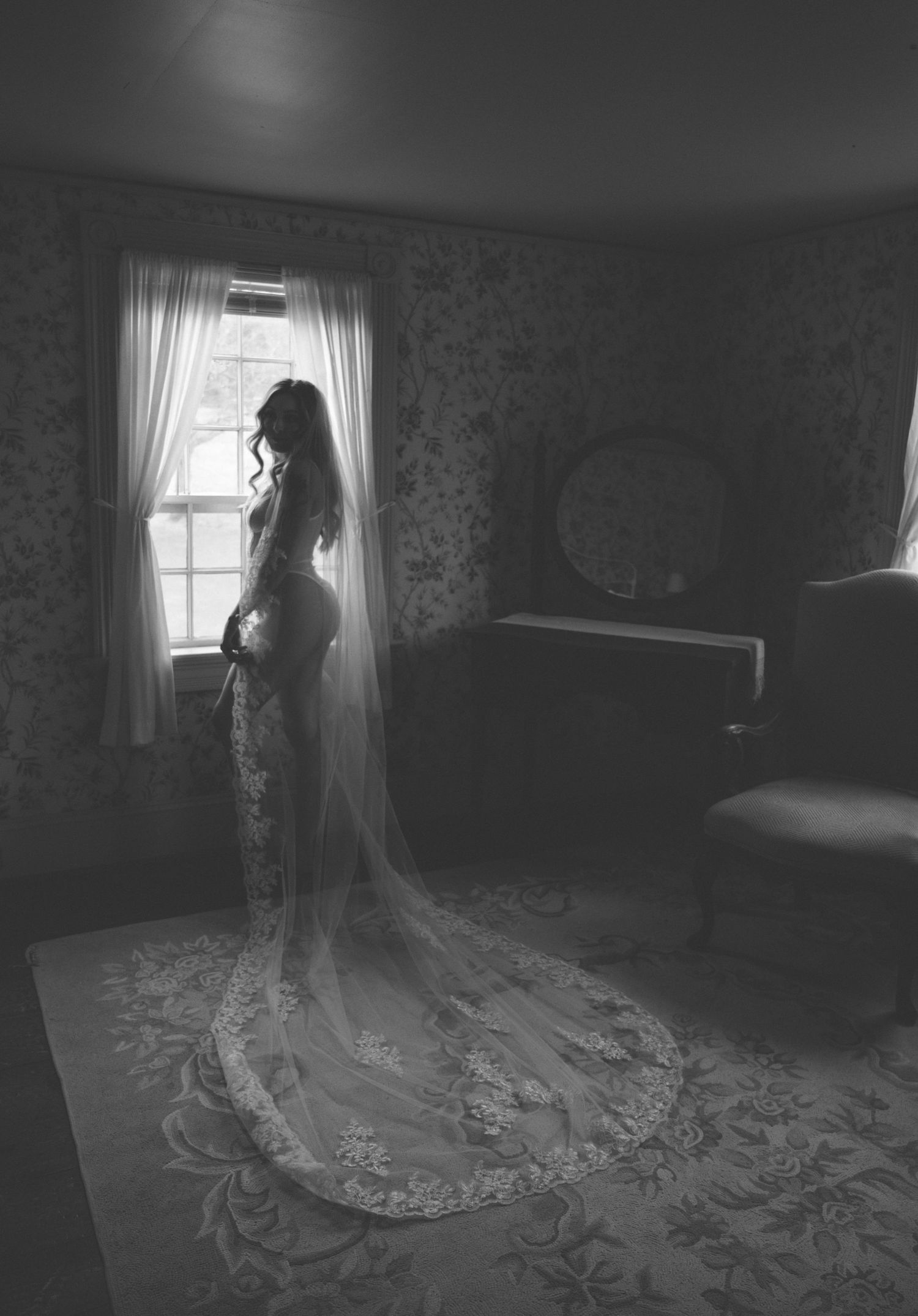 A silhouette of a bride in a lace bodysuit stands by a window in dramatic lighting.