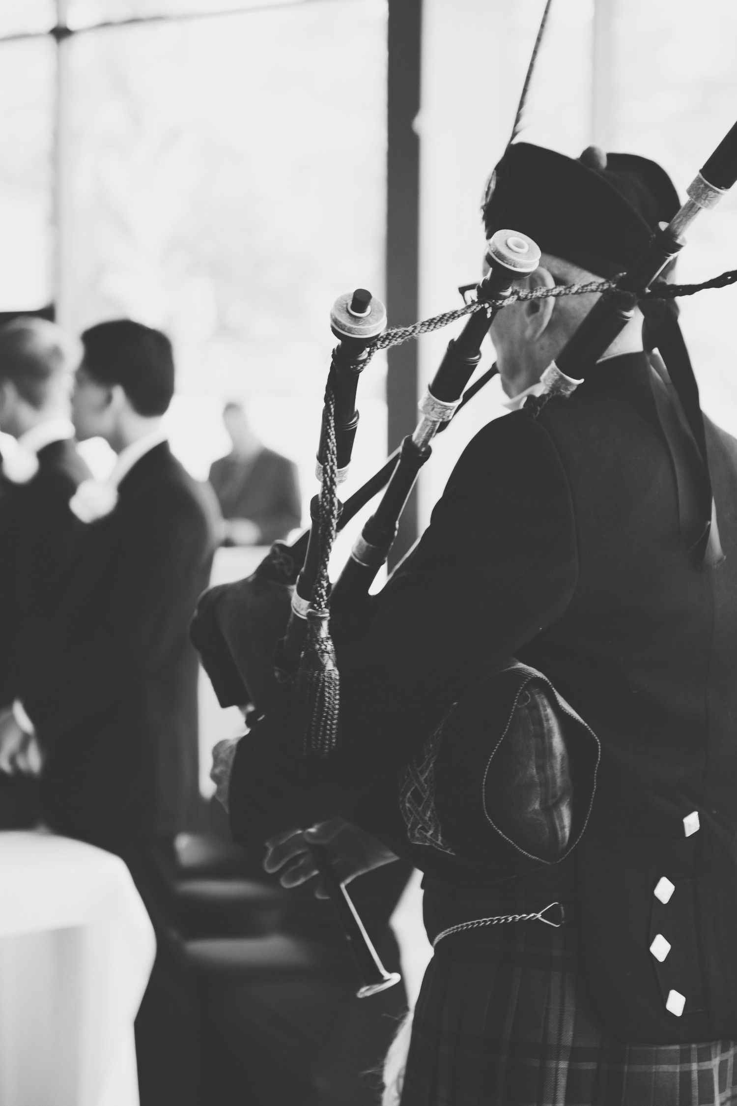 A bagpiper in traditional Scottish attire performs at an indoor event with guests mingling in the background.