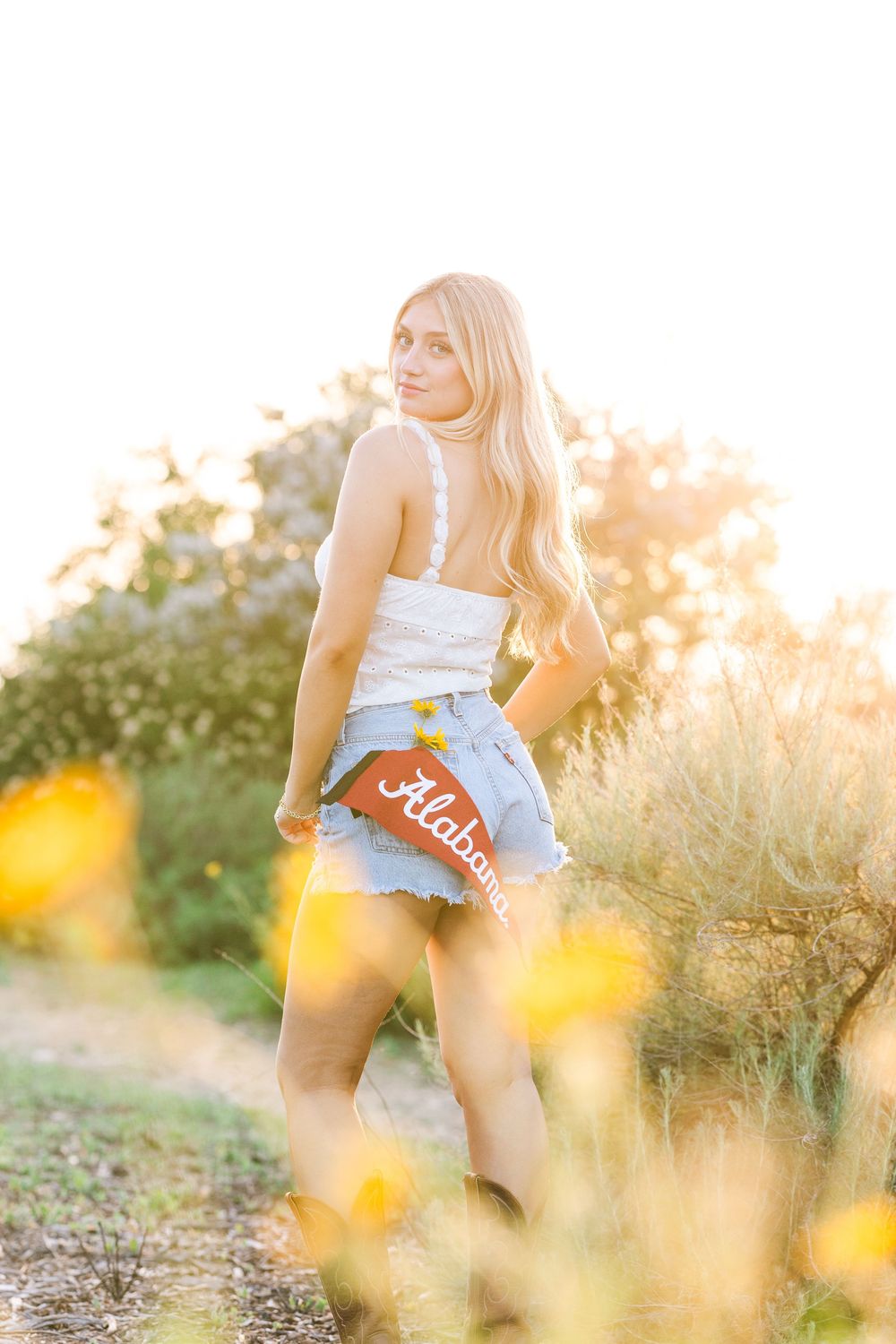 Woman sitting in golden wildflowers at sunset wearing denim shorts and white top in hazy summer sunlight.