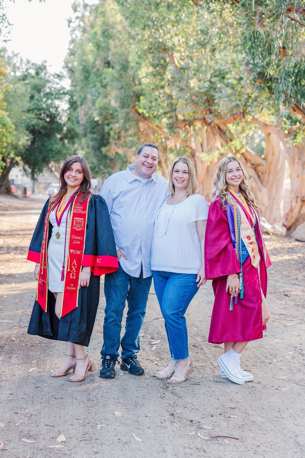 A family group poses outdoors on a tree-lined dirt path, with two wearing graduation gowns and stoles.