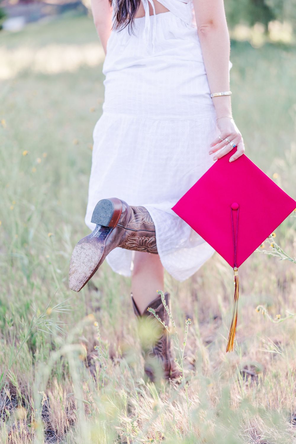 Graduate in white dress holds pink graduation cap and brown cowboy boots while standing in rustic field.