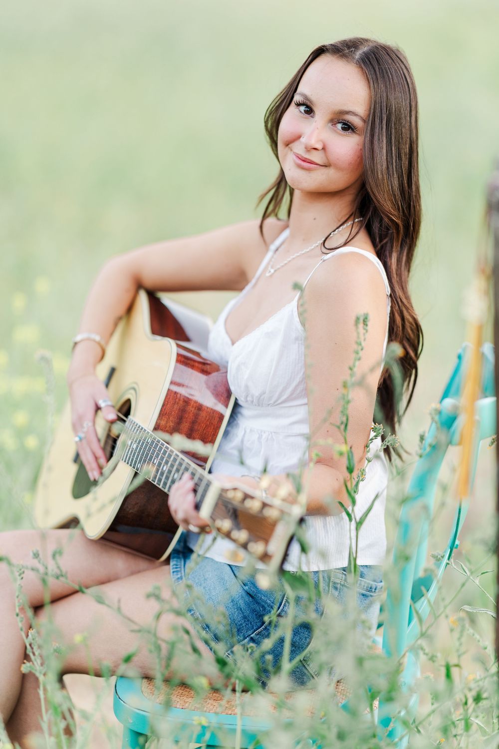 A musician in a white sundress plays acoustic guitar while sitting in a wildflower meadow on a summer day.